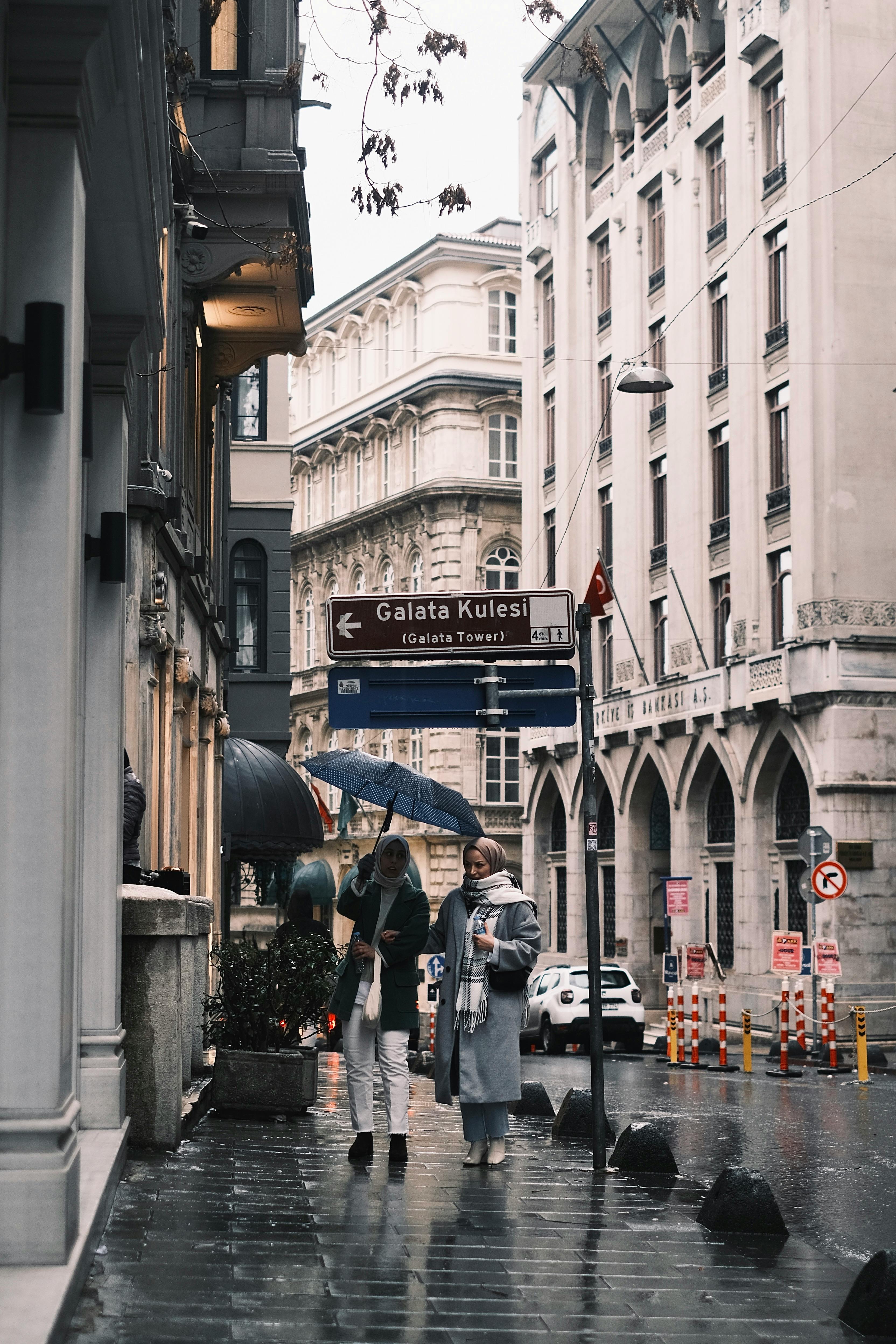 People walking with umbrellas near Galata Tower sign in Istanbul, capturing urban life in the rain.