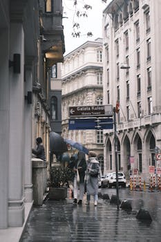 Pedestrians with umbrellas on a rainy day in Istanbul, near the historic Galata Tower, surrounded by classic architecture.