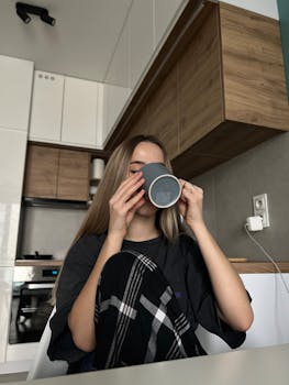 A young woman in a cozy kitchen drinks coffee in the morning, wearing casual attire.