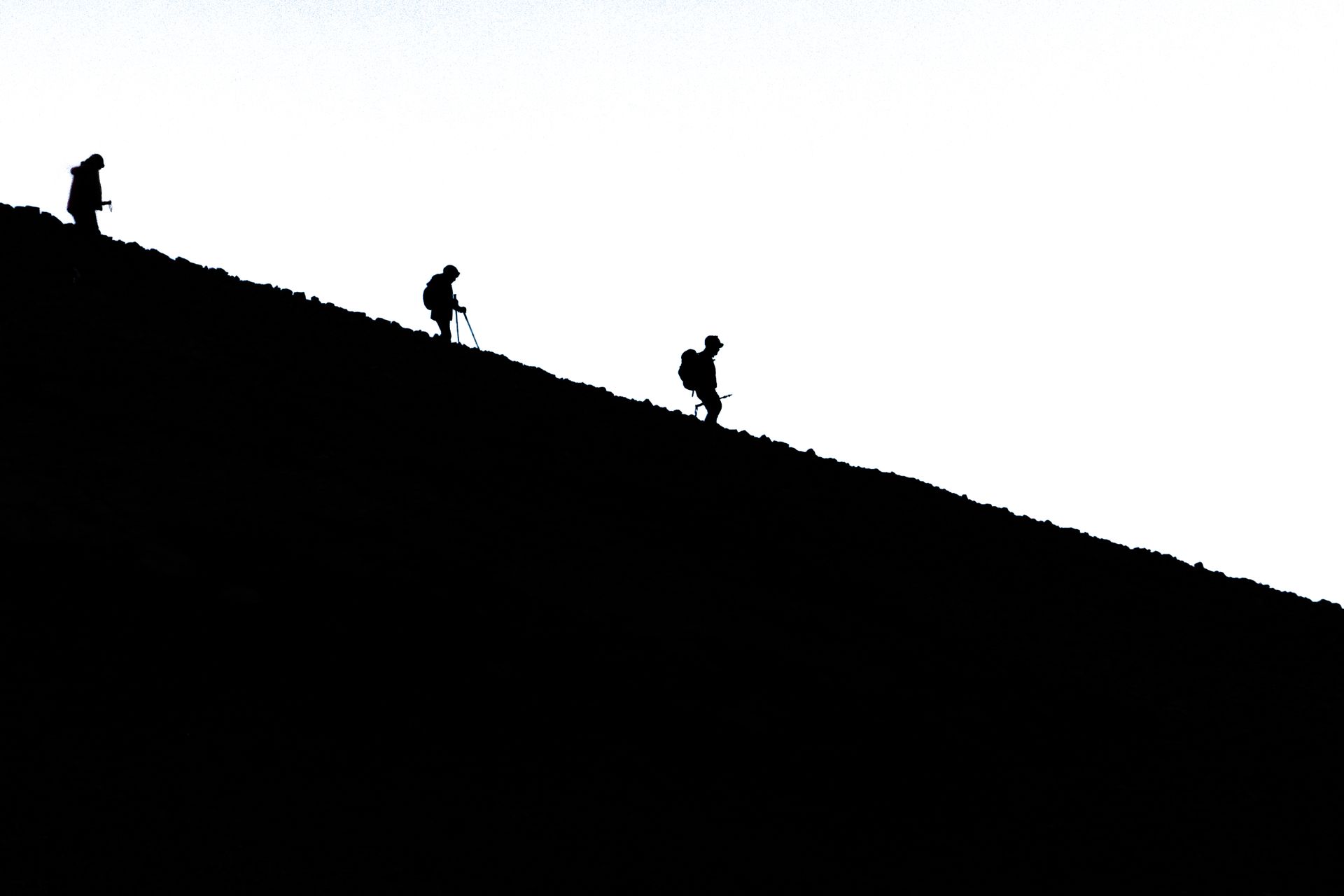 Silhouetted hikers trek downhill on a steep mountain slope, creating a dramatic black and white image.