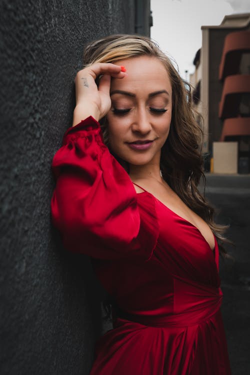 Young Woman in a Red Dress Standing with Her Back against the Wall of a Building in City 