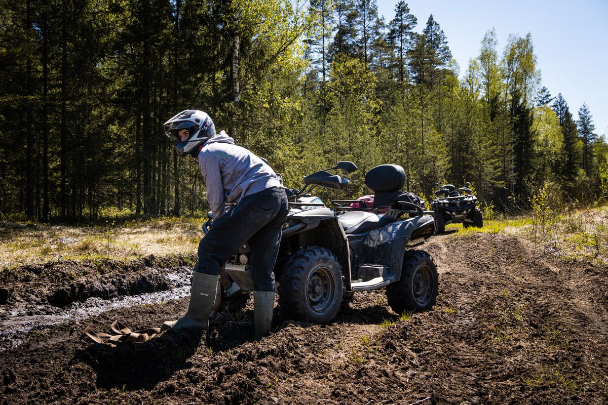 ATV rider in full safety gear including helmet and boots dealing with quad stuck in deep mud on forest trail