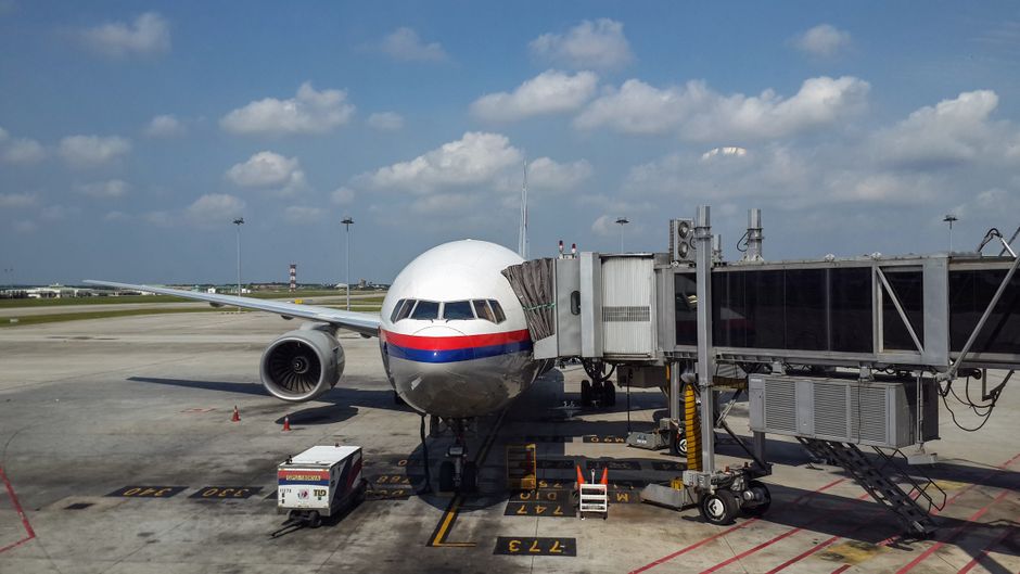 A commercial airplane parked at Kuala Lumpur airport, connected to a passenger boarding bridge under a clear blue sky.