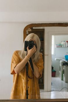 A blonde woman capturing a mirror selfie indoors wearing a mustard yellow shirt.