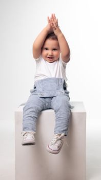Cute child wearing blue overalls and white shoes, clapping with joy in a studio shoot.