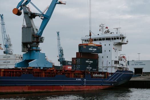 A cargo ship loaded with shipping containers at an industrial harbor with cranes.