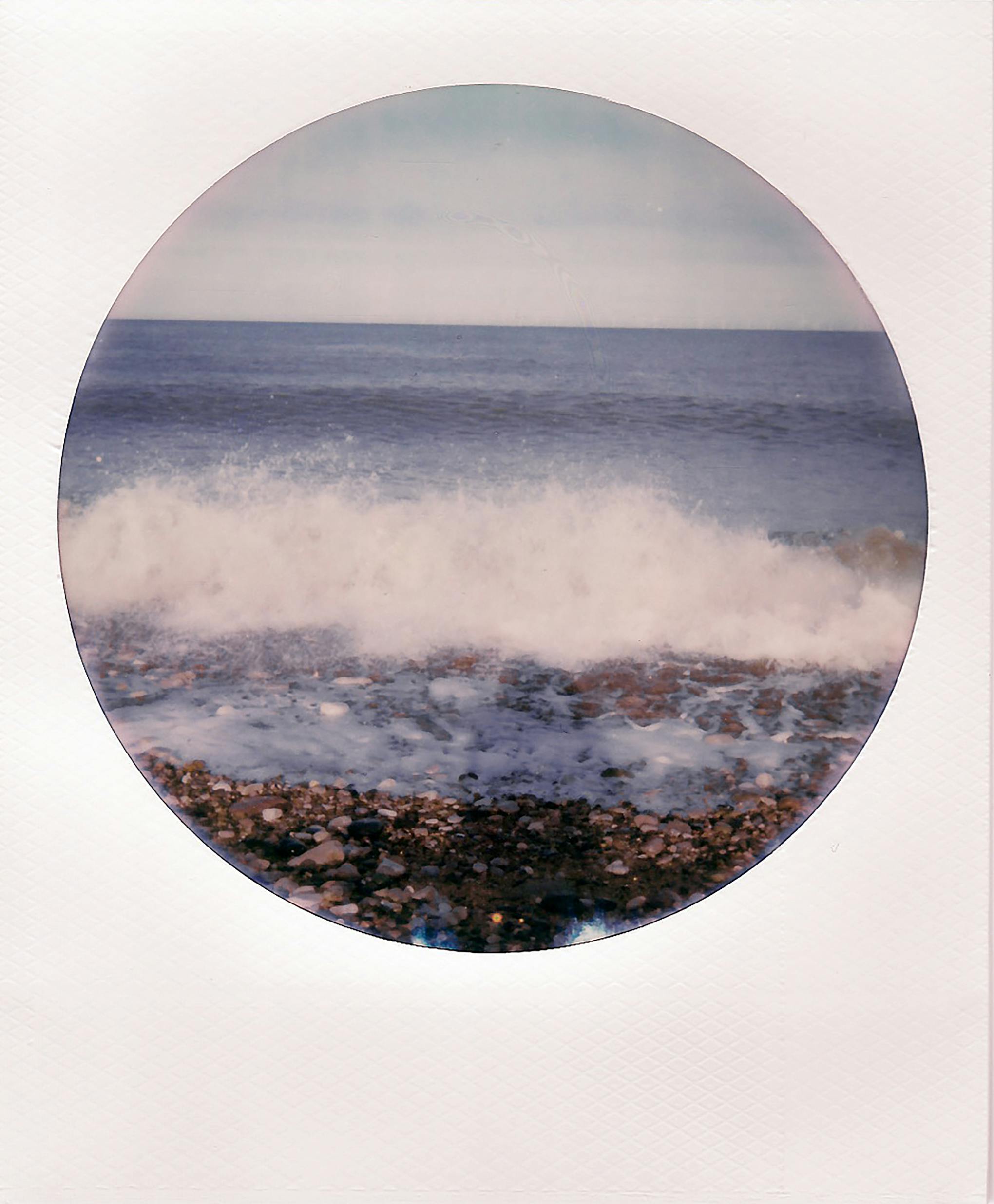 Vintage circular polaroid capturing waves crashing on a pebbled shore.