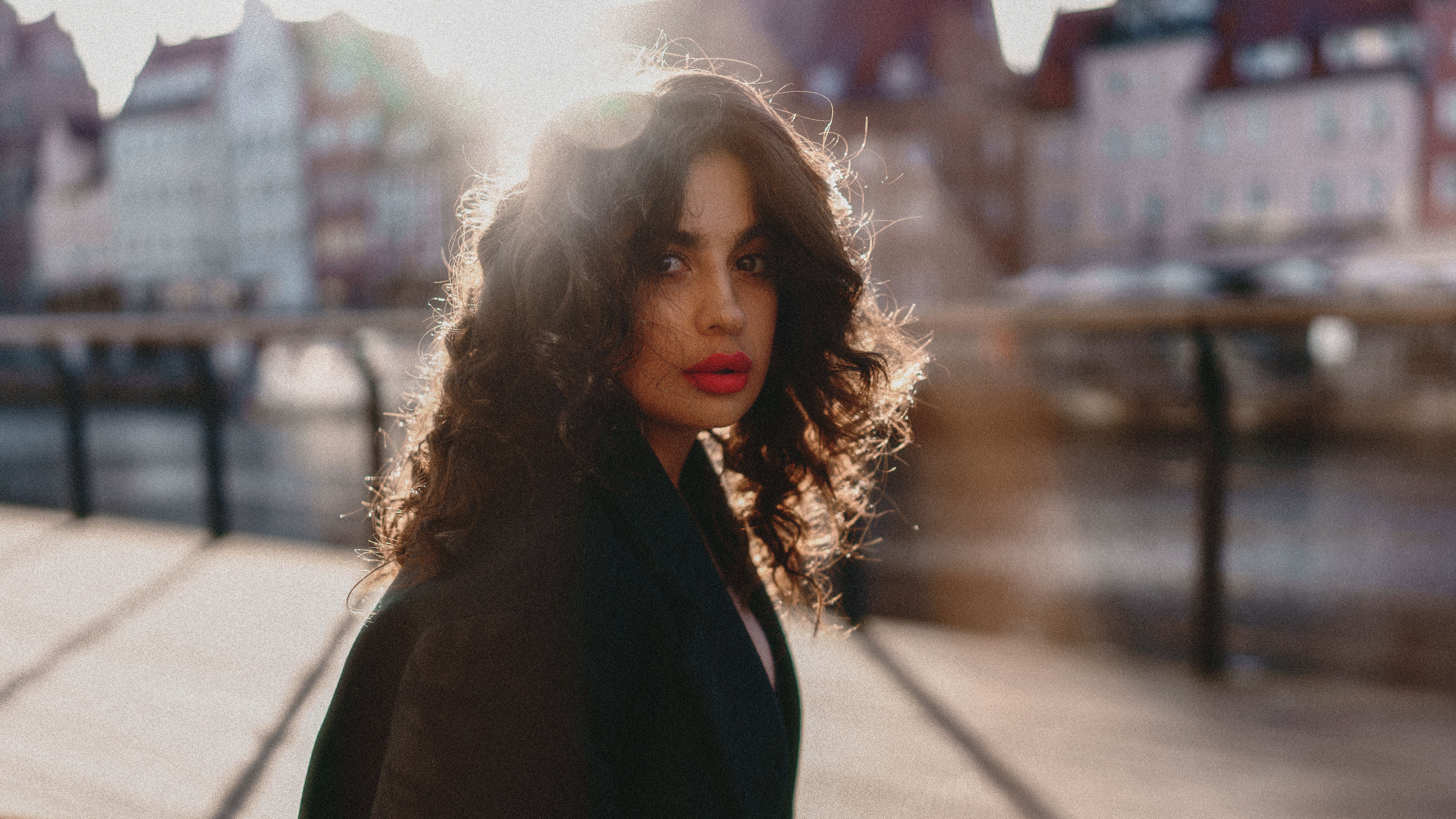 A stylish woman with curly hair and bold lips, captured in warm sunlight with a city backd