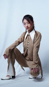 Portrait of an adult Asian woman kneeling in a textured brown outfit on a white background.