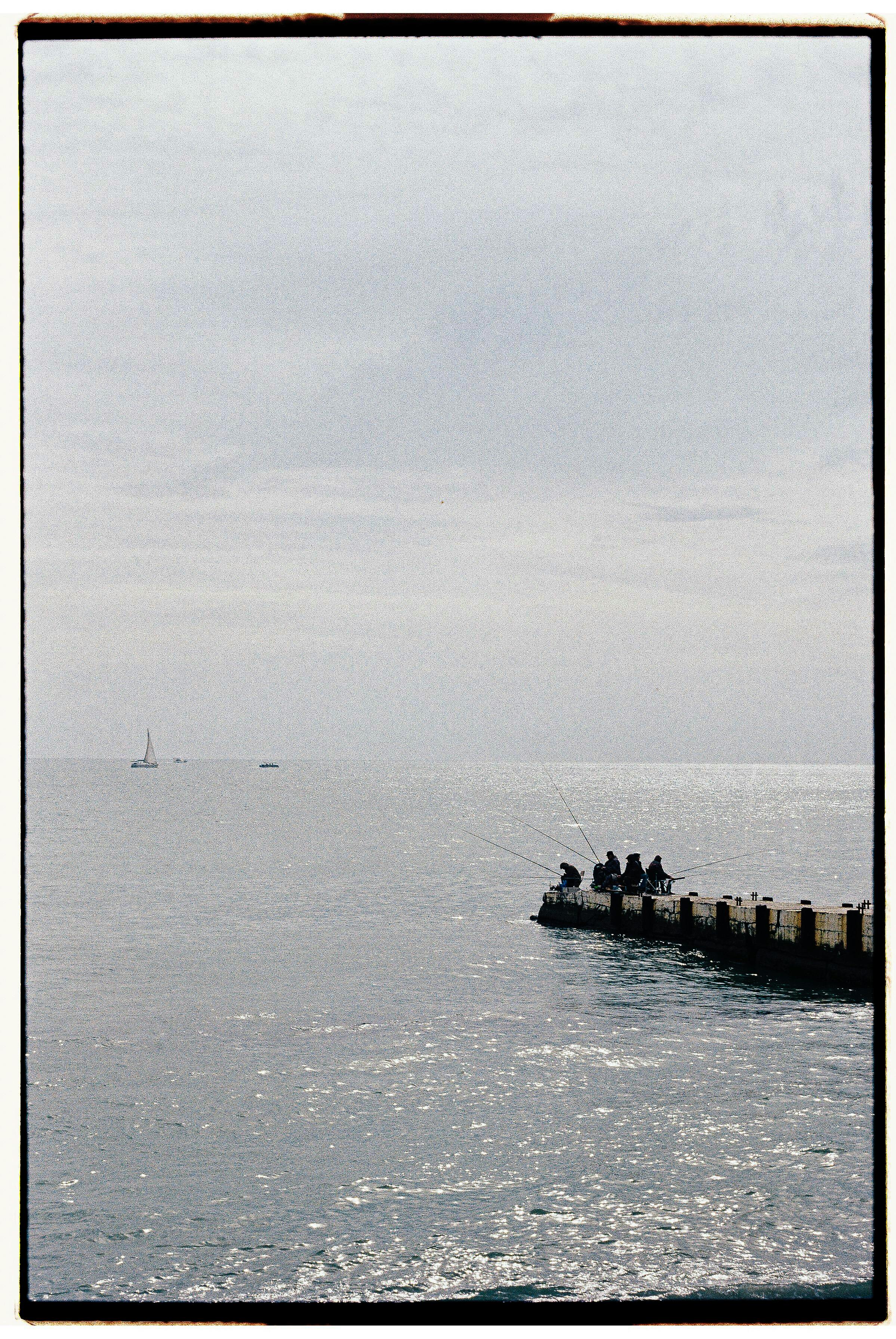 A tranquil scene of fishermen on a pier under an overcast sky, with distant sailboats.