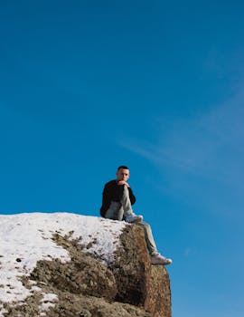 A man sits alone on a snowy cliff edge under a clear blue sky, capturing a moment of solitude and reflection.