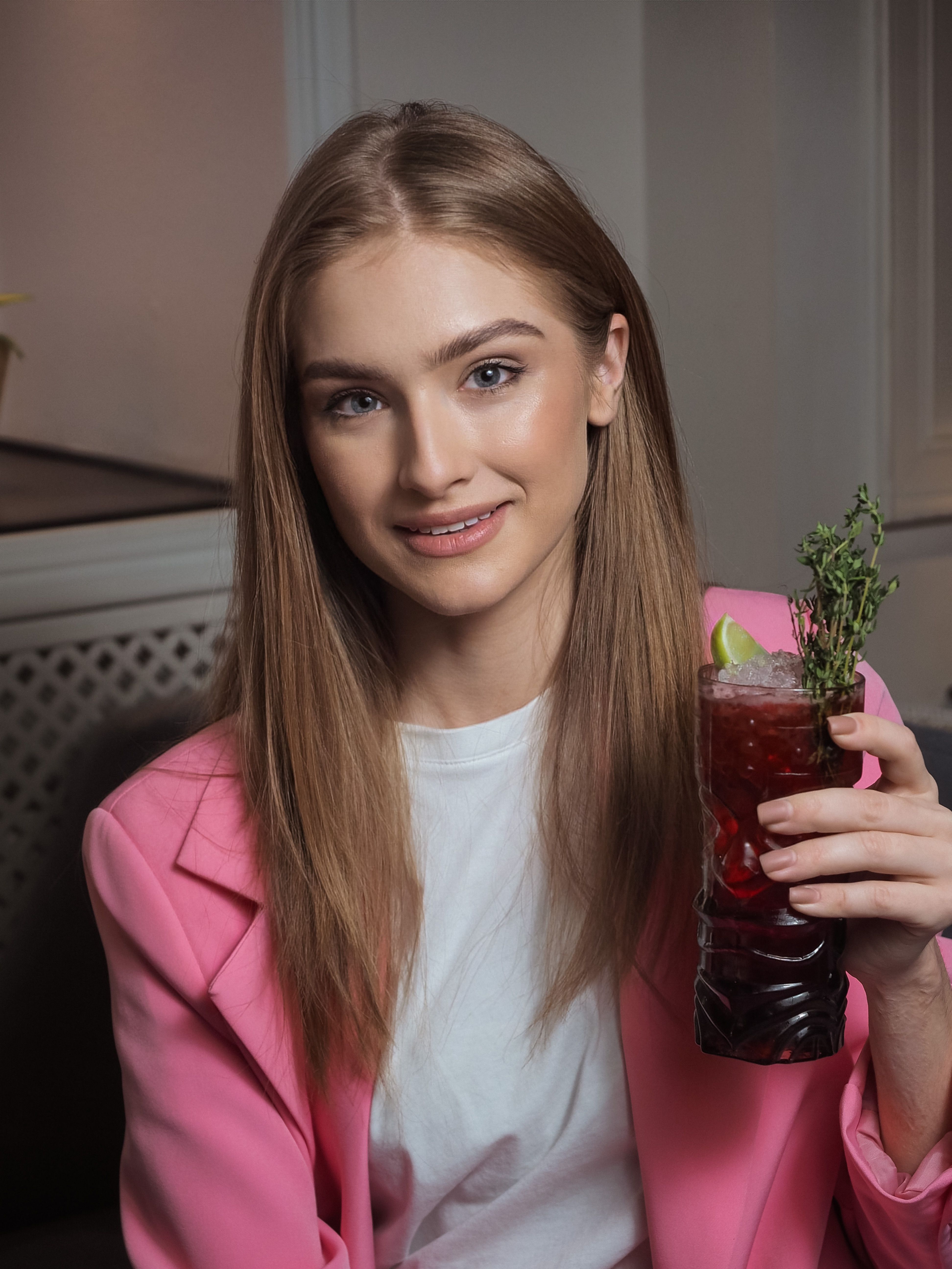 Young woman in a pink blazer smiling while holding a colorful cocktail indoors.