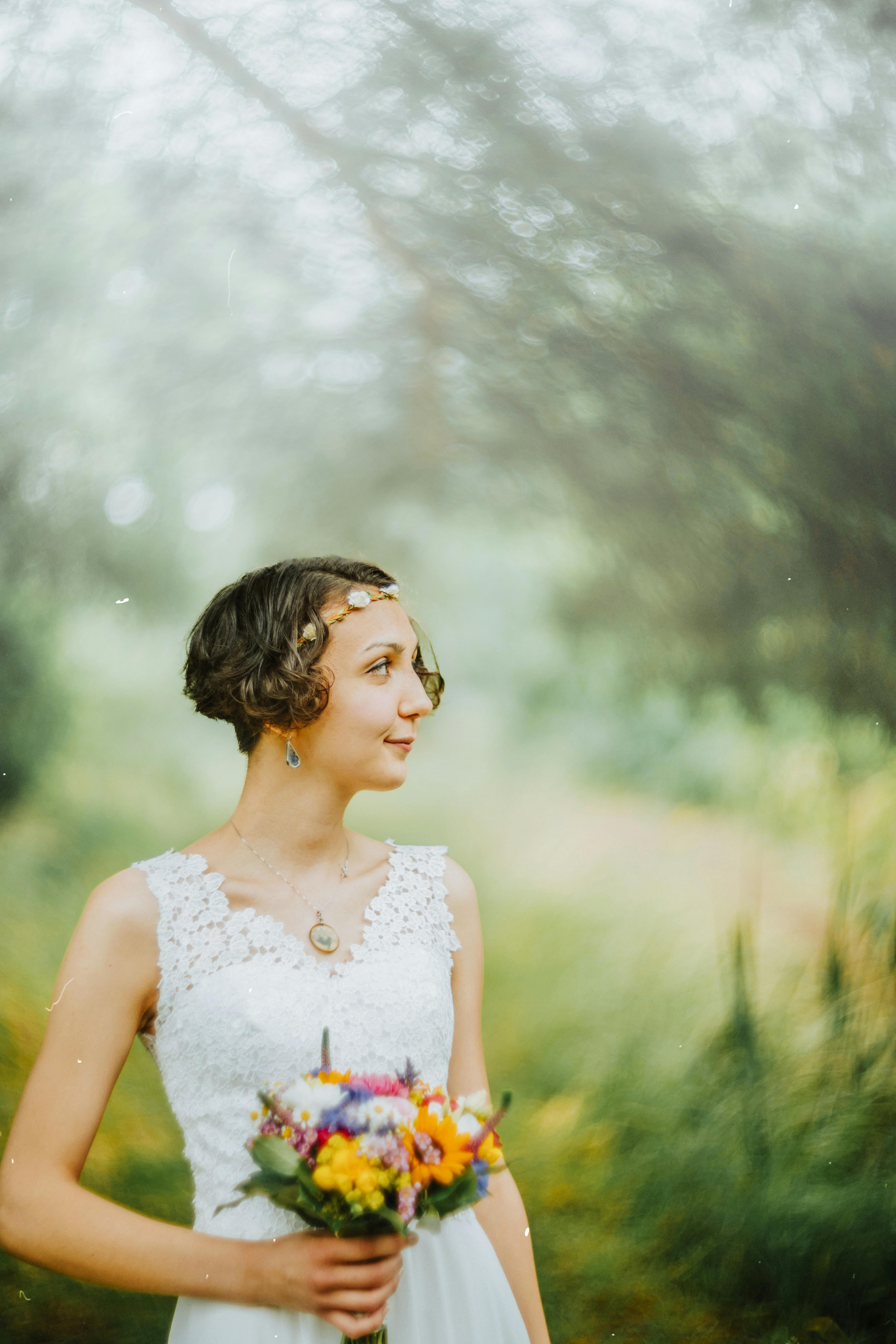A woman in a white dress with a short haircut holding a colorful bouquet in a misty, outdoor setting.
