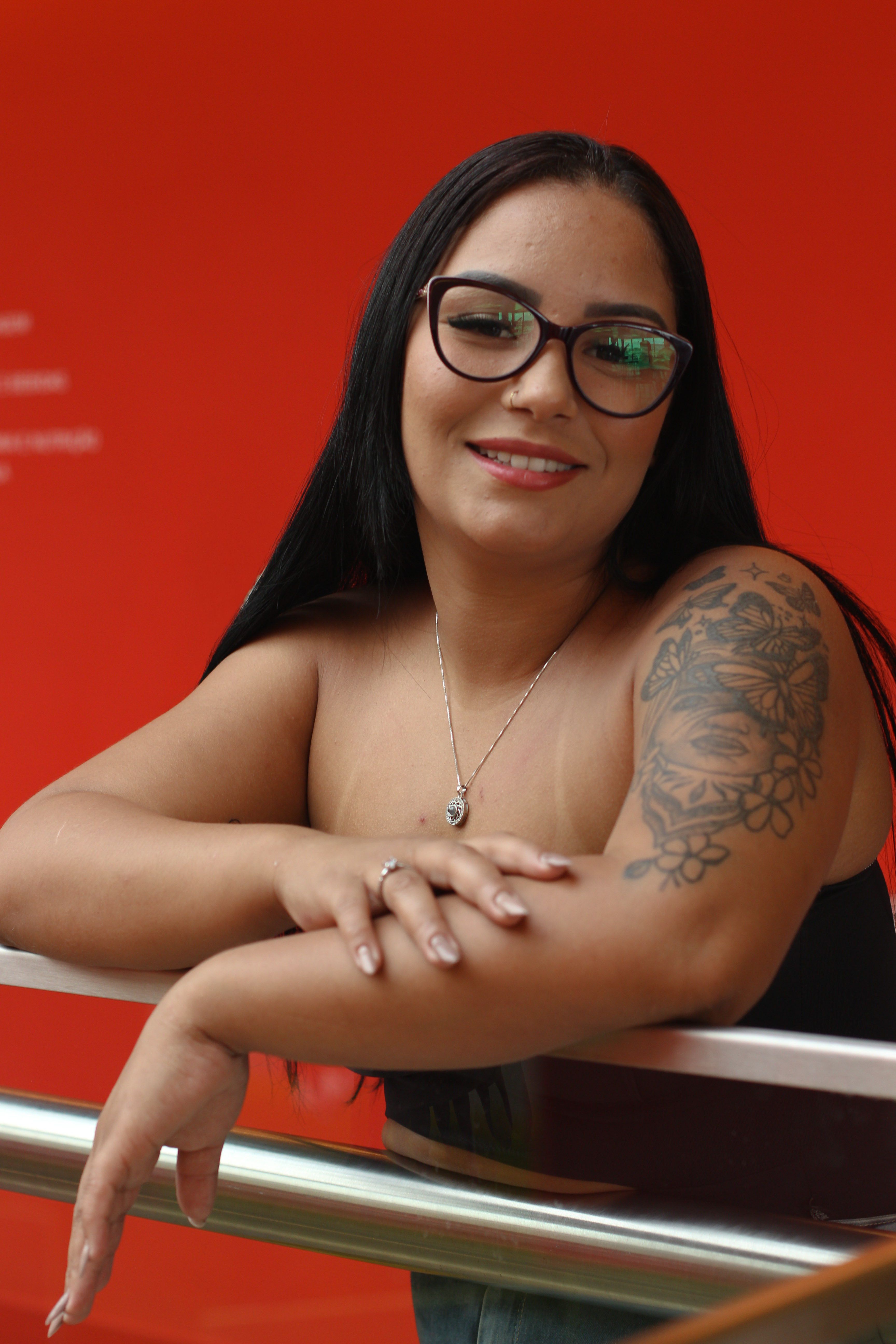 Confident woman smiling with floral tattoo, black hair, and glasses against a bold red backdrop.