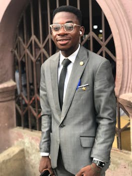 Young man in a gray suit smiling confidently, standing outdoors with eyeglasses and airpods.