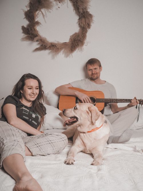 Free A man plays guitar while a woman and dog relax on a bed in a cozy indoor setting. Stock Photo