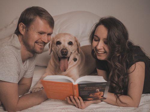 Free Smiling couple with their dog enjoying a book together in a cozy and relaxed bedroom setting. Stock Photo