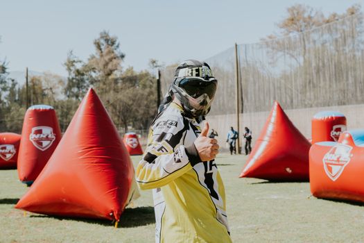 A paintball player in full gear and goggles giving a thumbs up during a game outdoors on a sunny day.