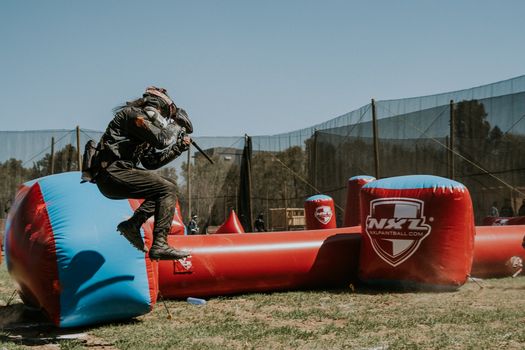Dynamic paintball match action with player jumping over inflatable barriers in an outdoor field.