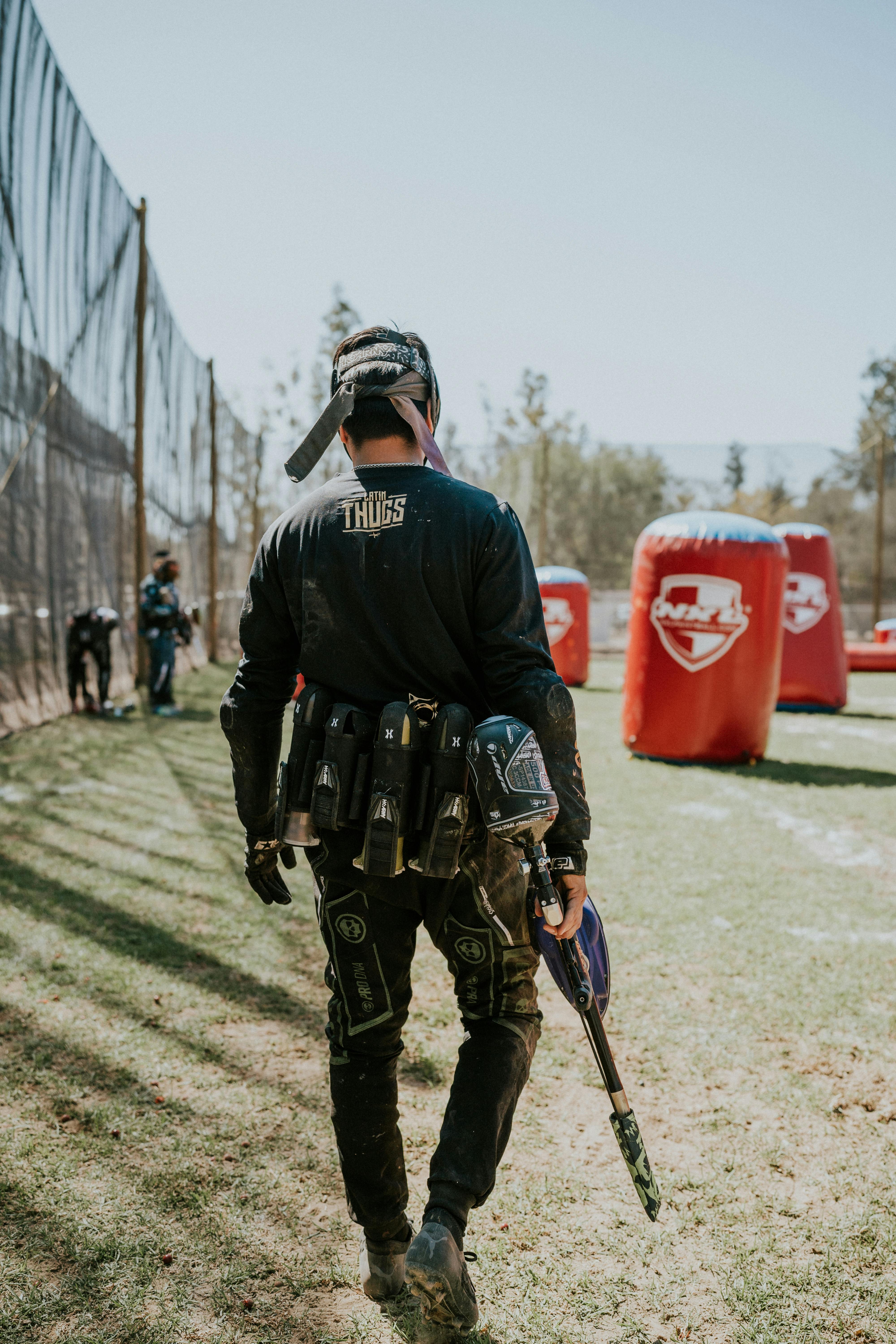 Ein Mann geht mit seiner Ausrüstung in der Hand im hellen Sonnenlicht über ein Paintballfeld im Freien. Prescription Goggle Inserts - A man walks through an outdoor paintball field under bright sunlight, holding equipment.