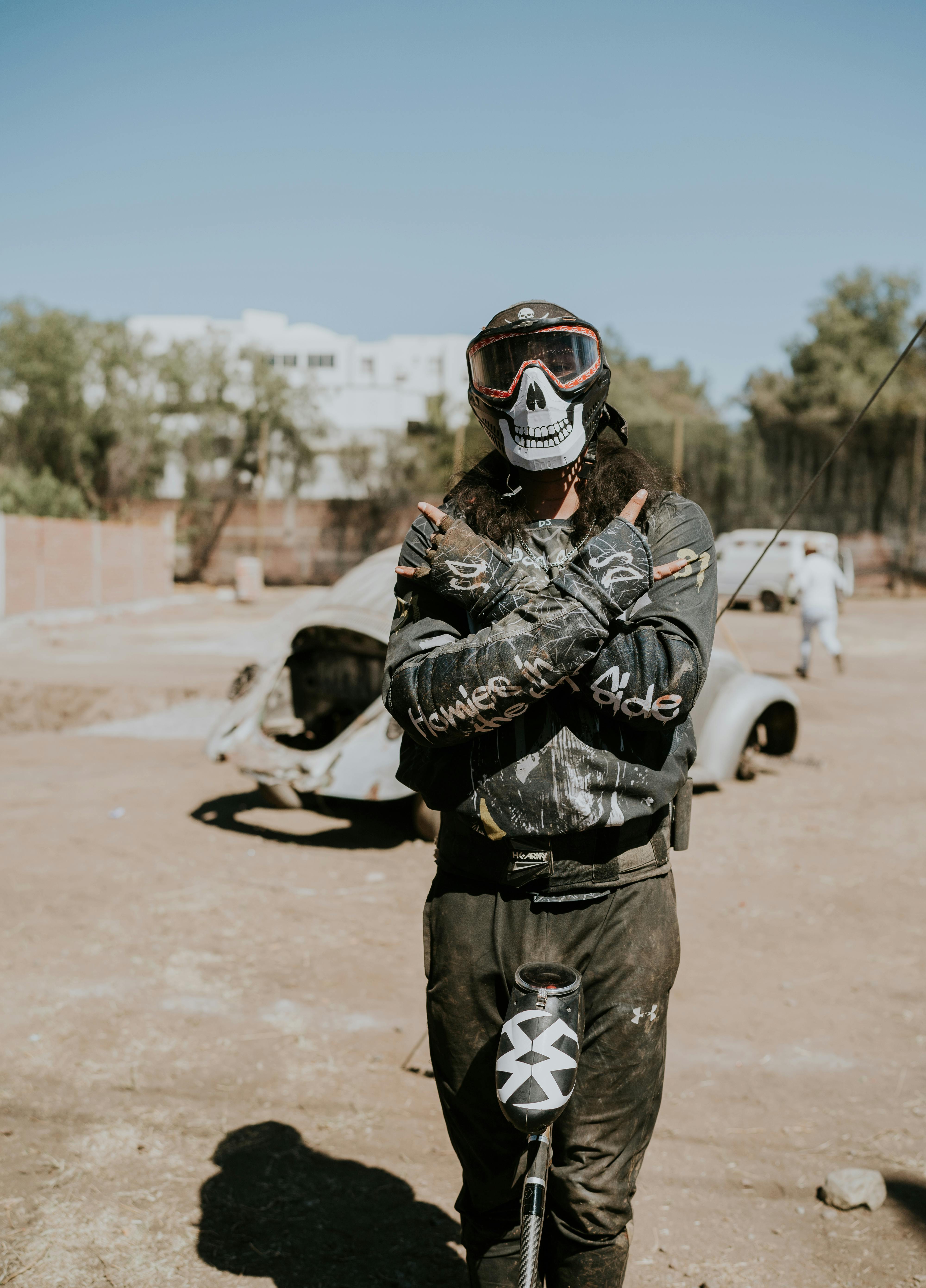 Un joueur de paintball avec un masque de crâne se tient avec confiance à l'extérieur, les bras croisés. Prescription Goggle Inserts - A paintball player with a skull mask stands confidently outdoors with arms crossed.