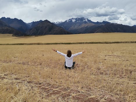 A woman sits in a field with arms raised, embracing freedom in a stunning mountain landscape.