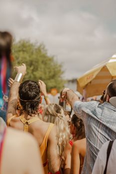 Back view of people at an outdoor festival with raised hands under cloudy sky.