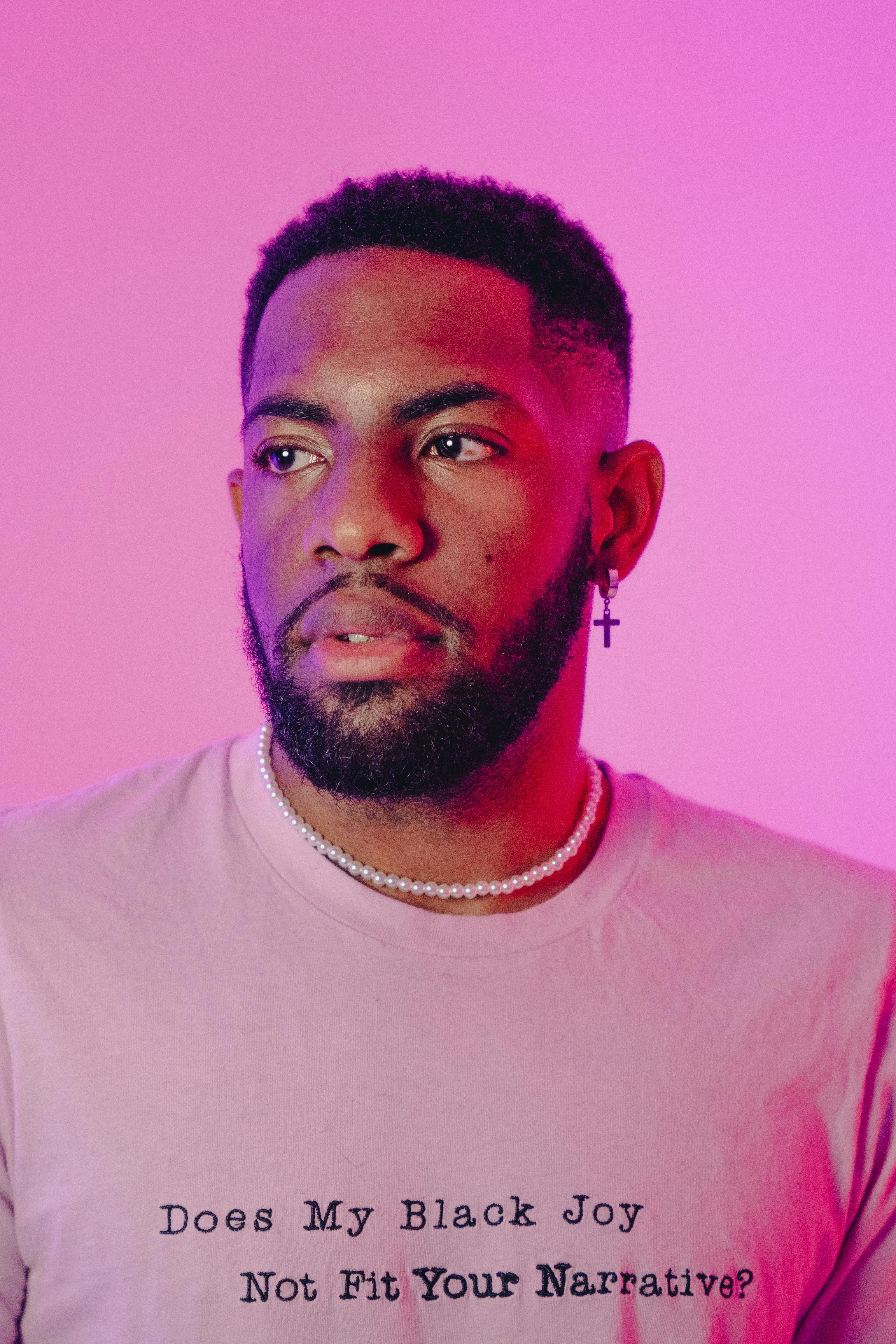 Portrait of a stylish young man with a beard and jewelry against a vibrant pink backdrop.