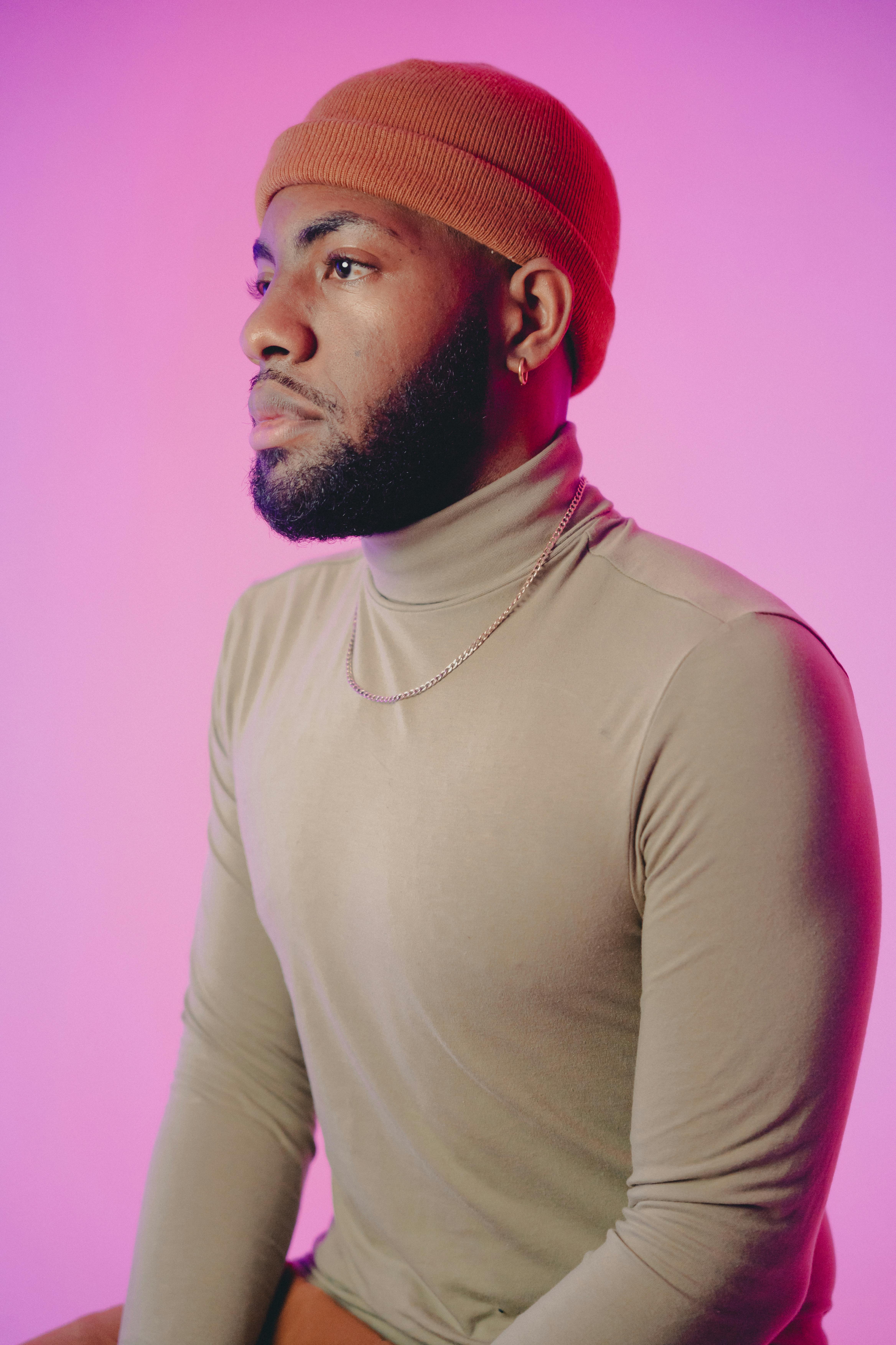 A fashionable man in a turtleneck sweater and knit hat poses against a vibrant pink background.