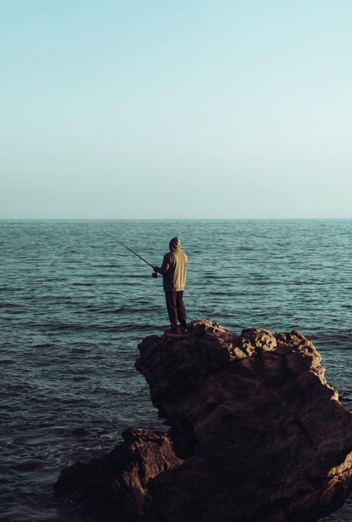 Free A lone fisherman casts his rod from a rocky outcrop overlooking the sea. Stock Photo