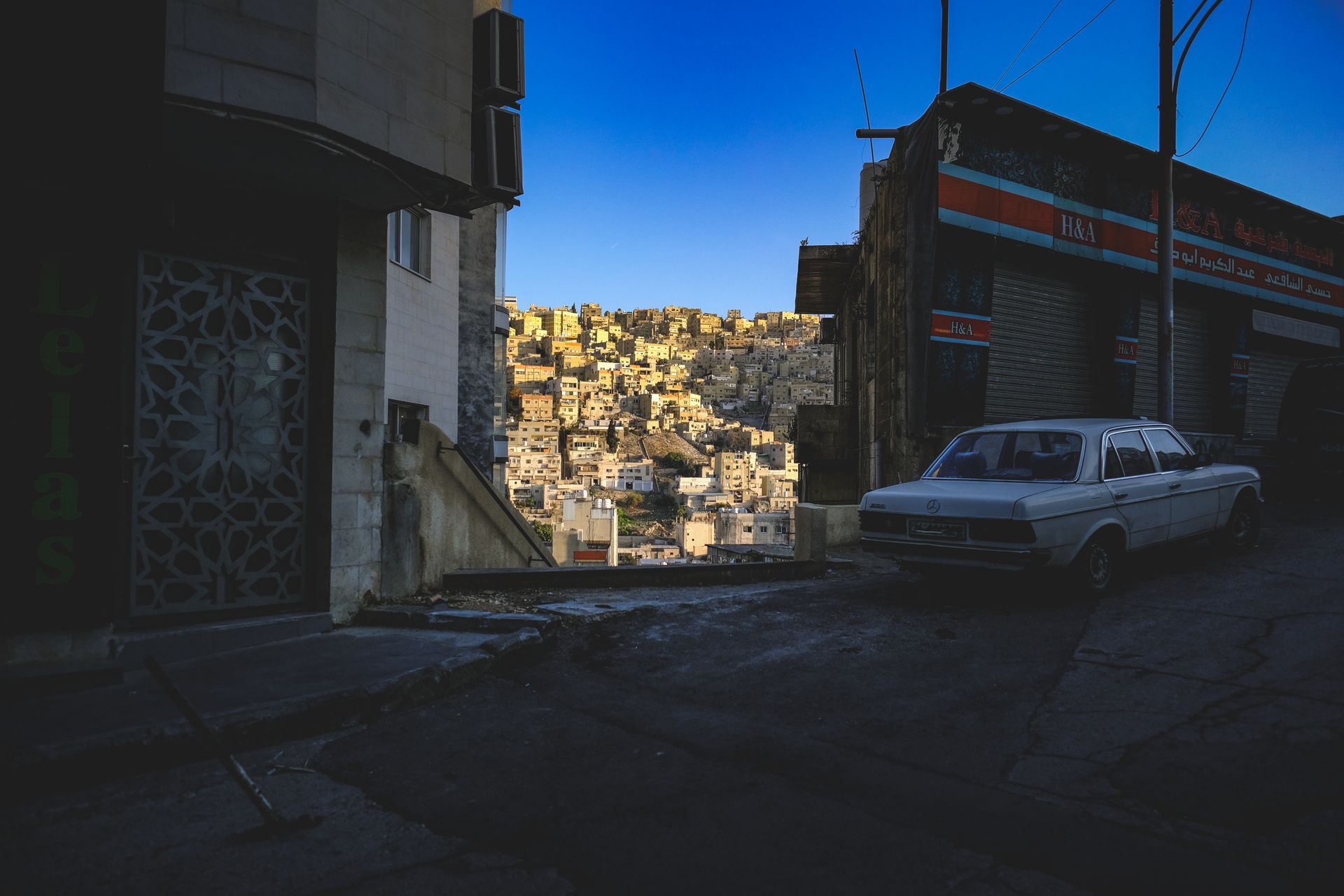 A contrasting cityscape featuring sunlit hillside houses viewed from a shadowed urban alley with a vintage car.
