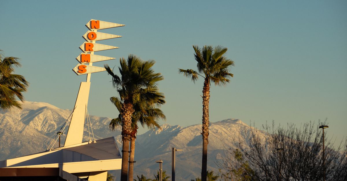 A retro Norms restaurant sign framed by palm trees and snow-capped mountains in Rialto, CA.