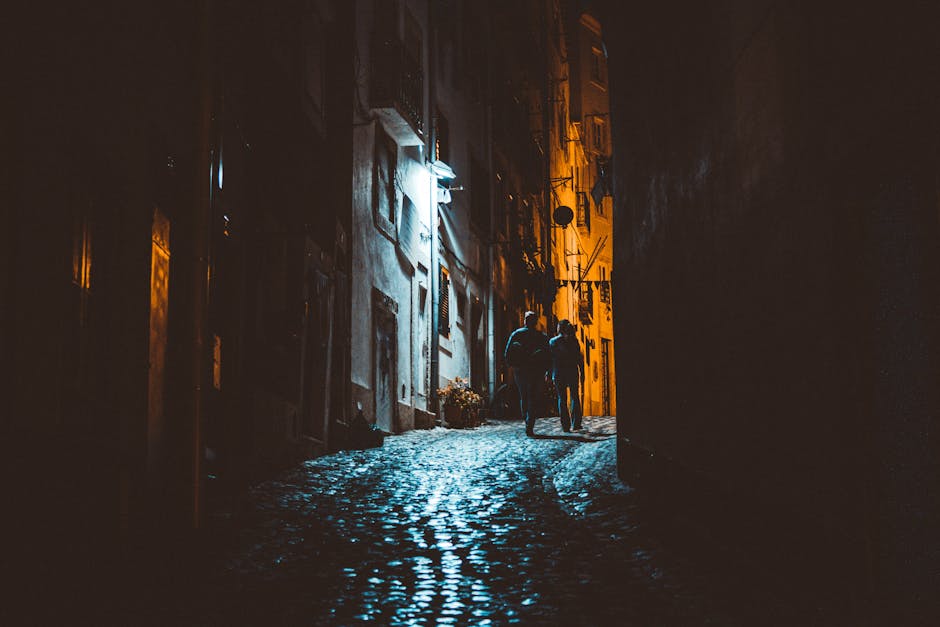 Silhouettes of people walking at night in a narrow cobblestone alley with dramatic lighting.