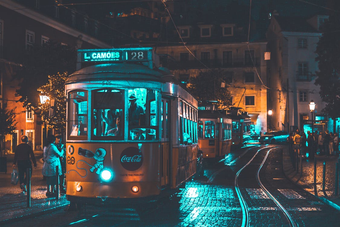 Vintage tram lit by blue and orange lights travels through a lively street in Lisbon at night.