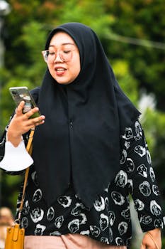 Portrait of an Asian woman wearing a hijab and eyeglasses, using a smartphone outdoors.