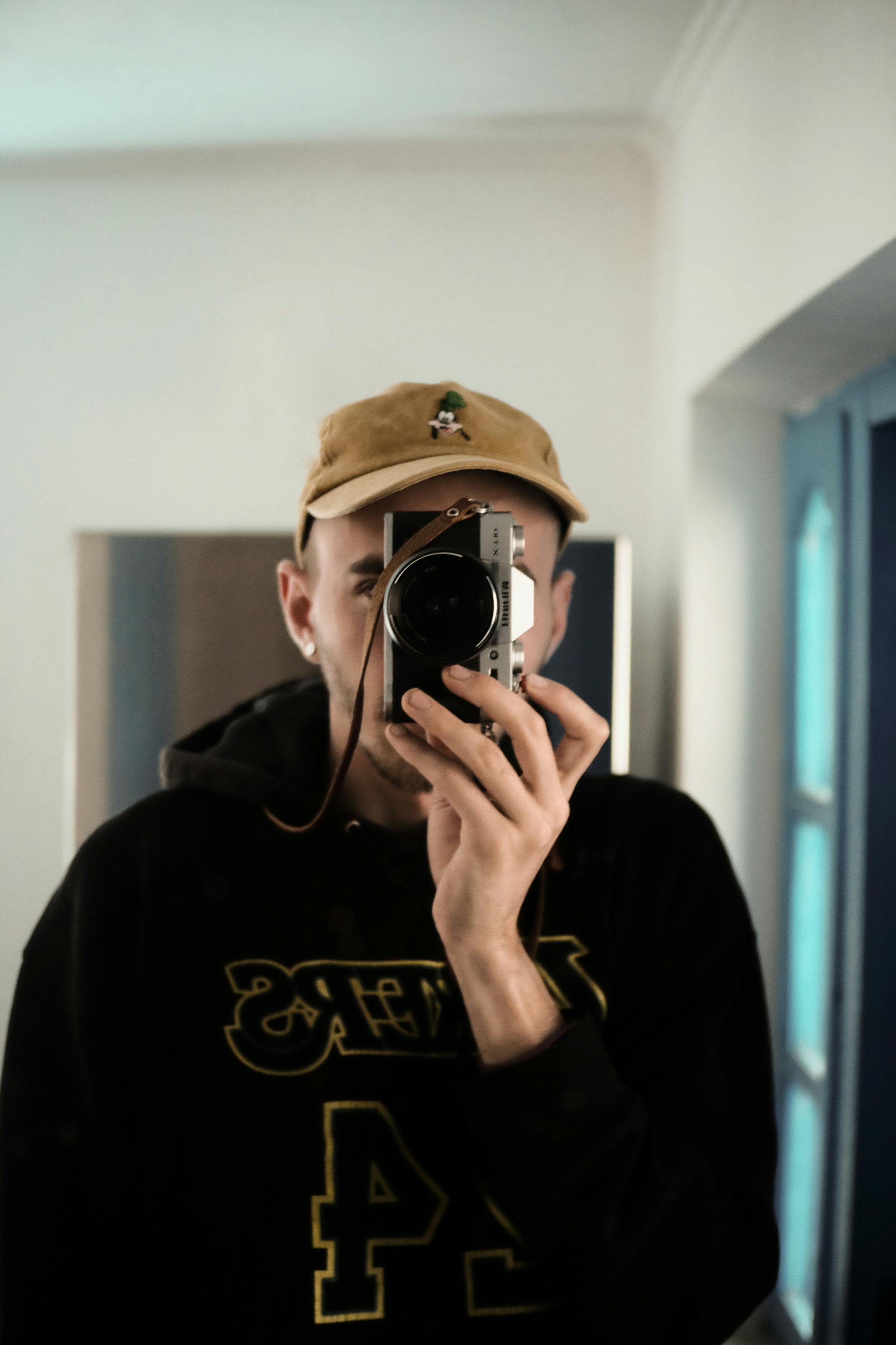 A young man wearing a cap and hoodie takes a selfie in a mirror with his camera indoors.