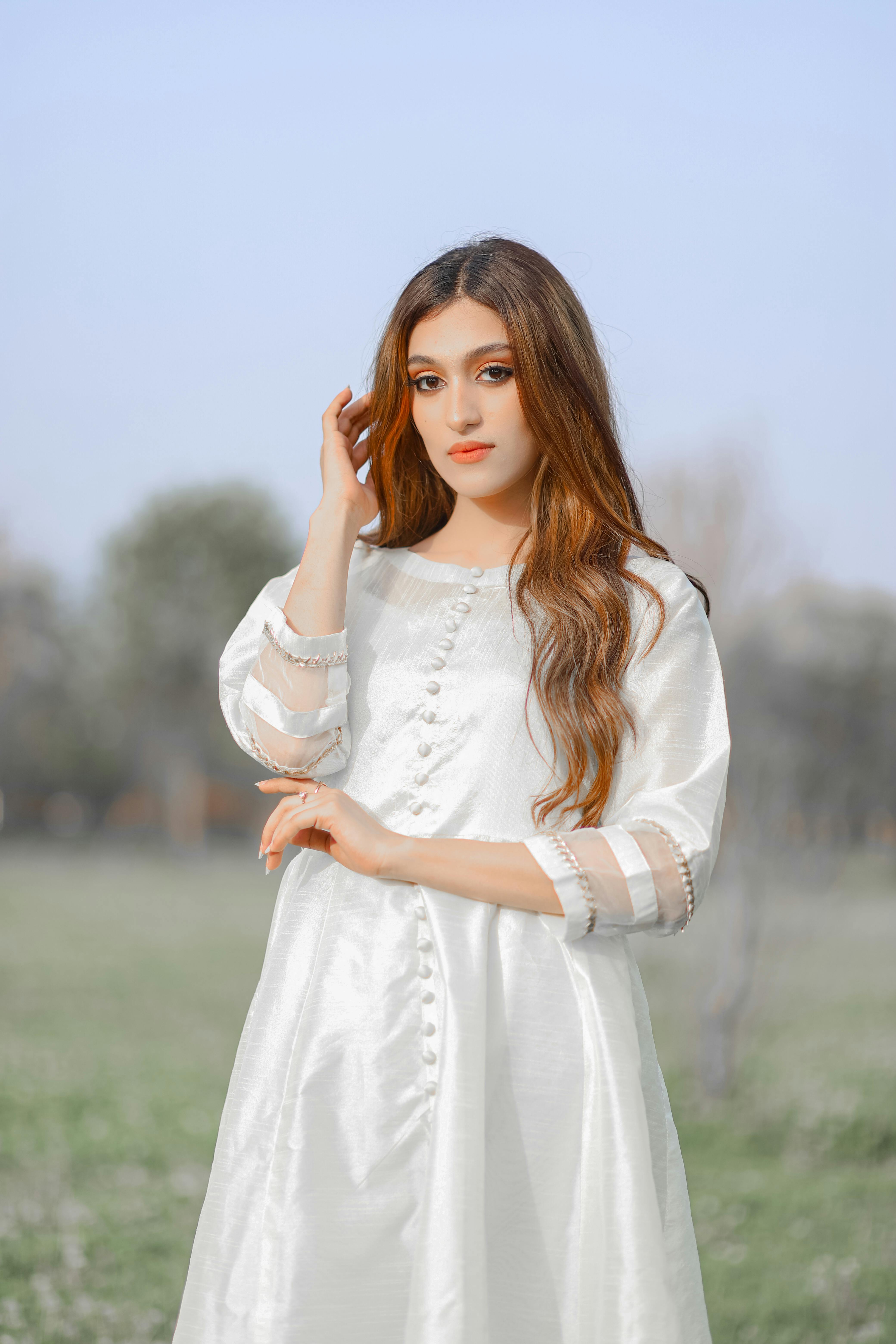 A graceful portrait of a woman in a white dress, standing outdoors in a serene setting.