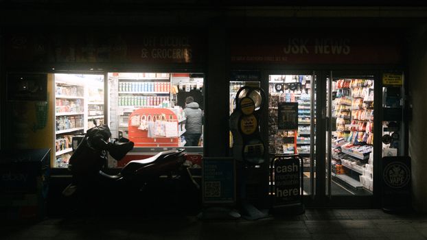 A dimly lit convenience store in London at night with a motorbike parked outside.