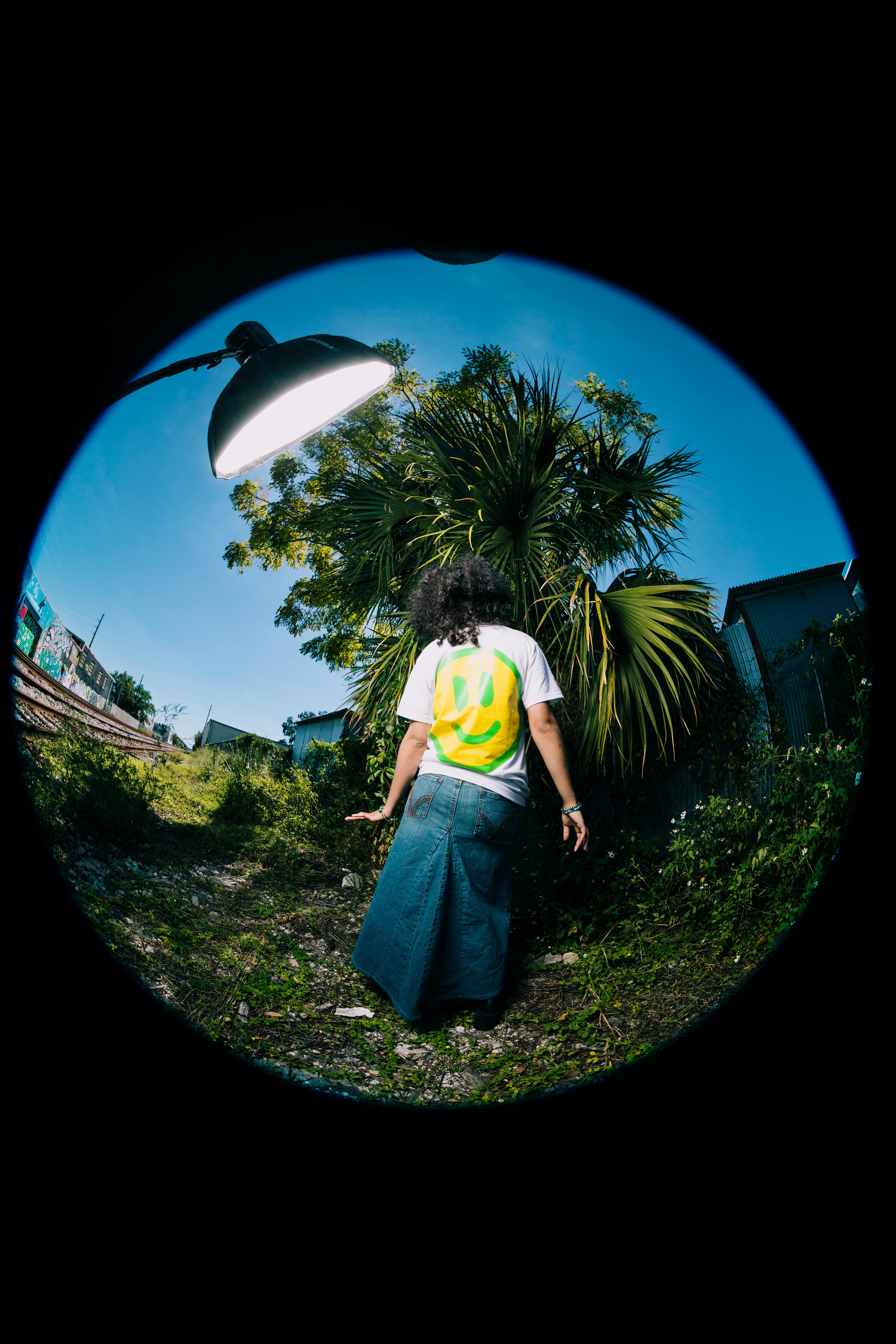 A person with dreadlocks poses with a smiley face t-shirt under urban streetlight.