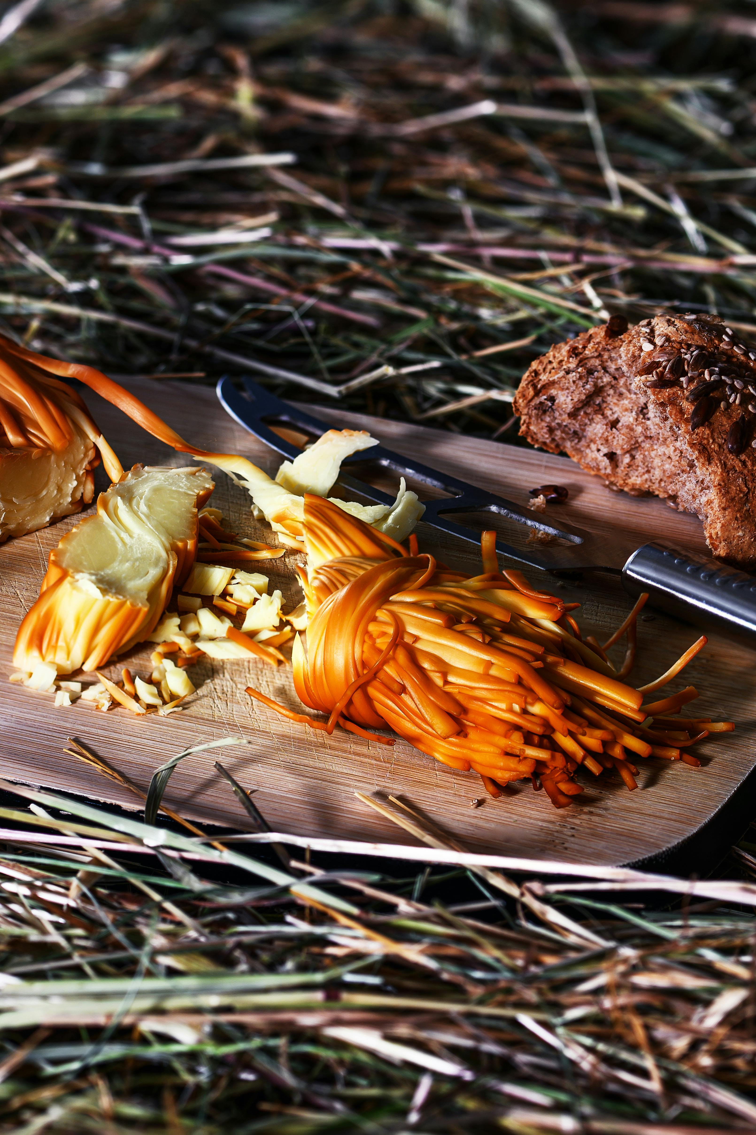 Close-up of cheese, knife, and bread on a wooden board over hay, emphasizing rustic charm.