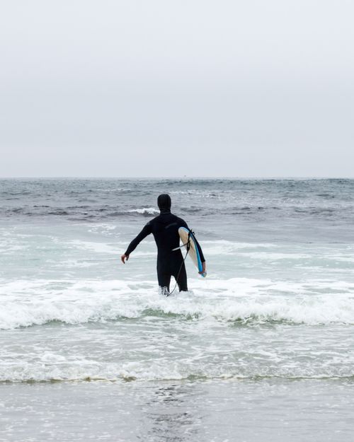 Free Surfer in wetsuit entering the ocean with a surfboard on an overcast day. Stock Photo