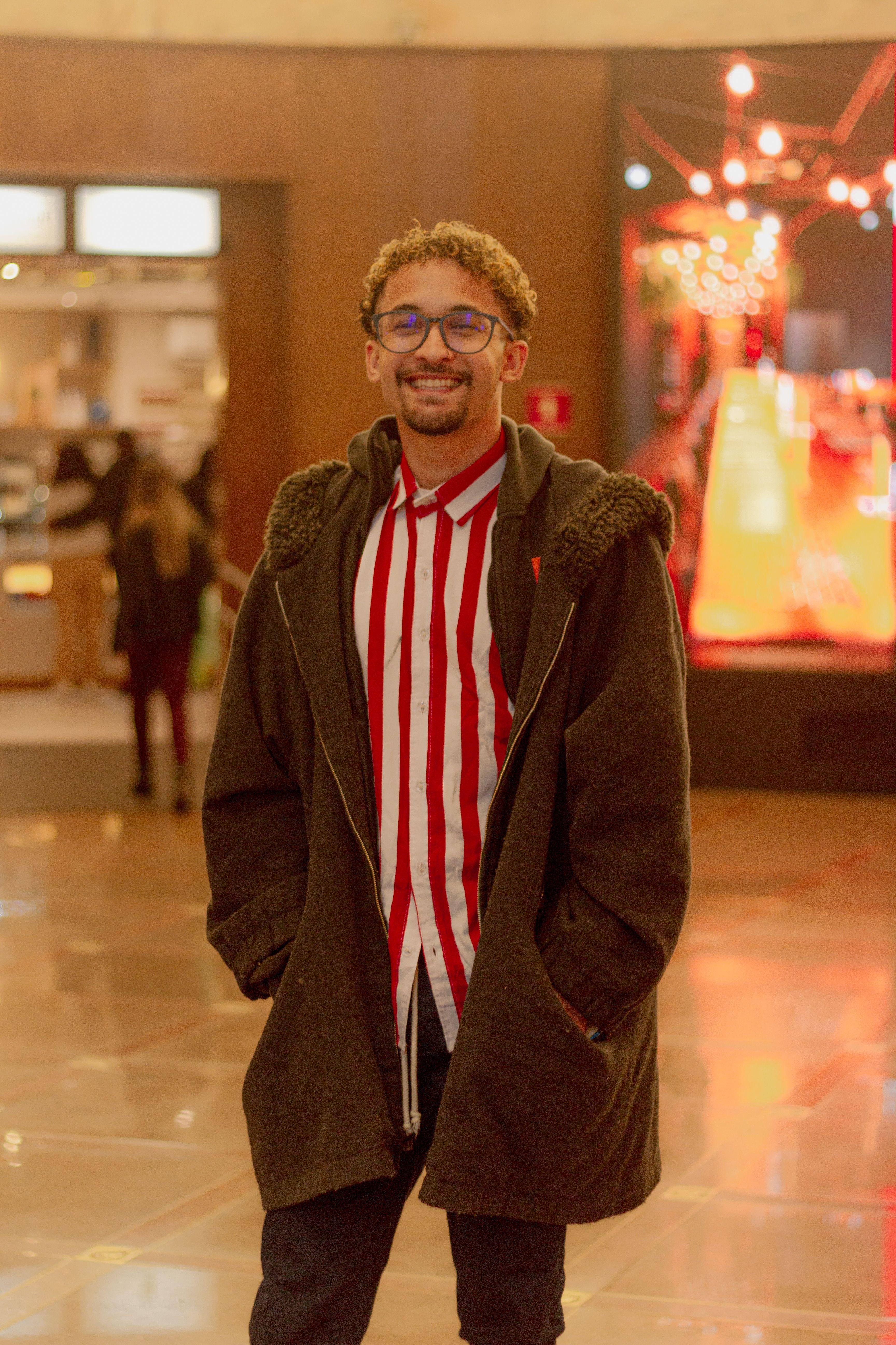 Young man in eyeglasses wearing a coat and striped shirt, smiling indoors.