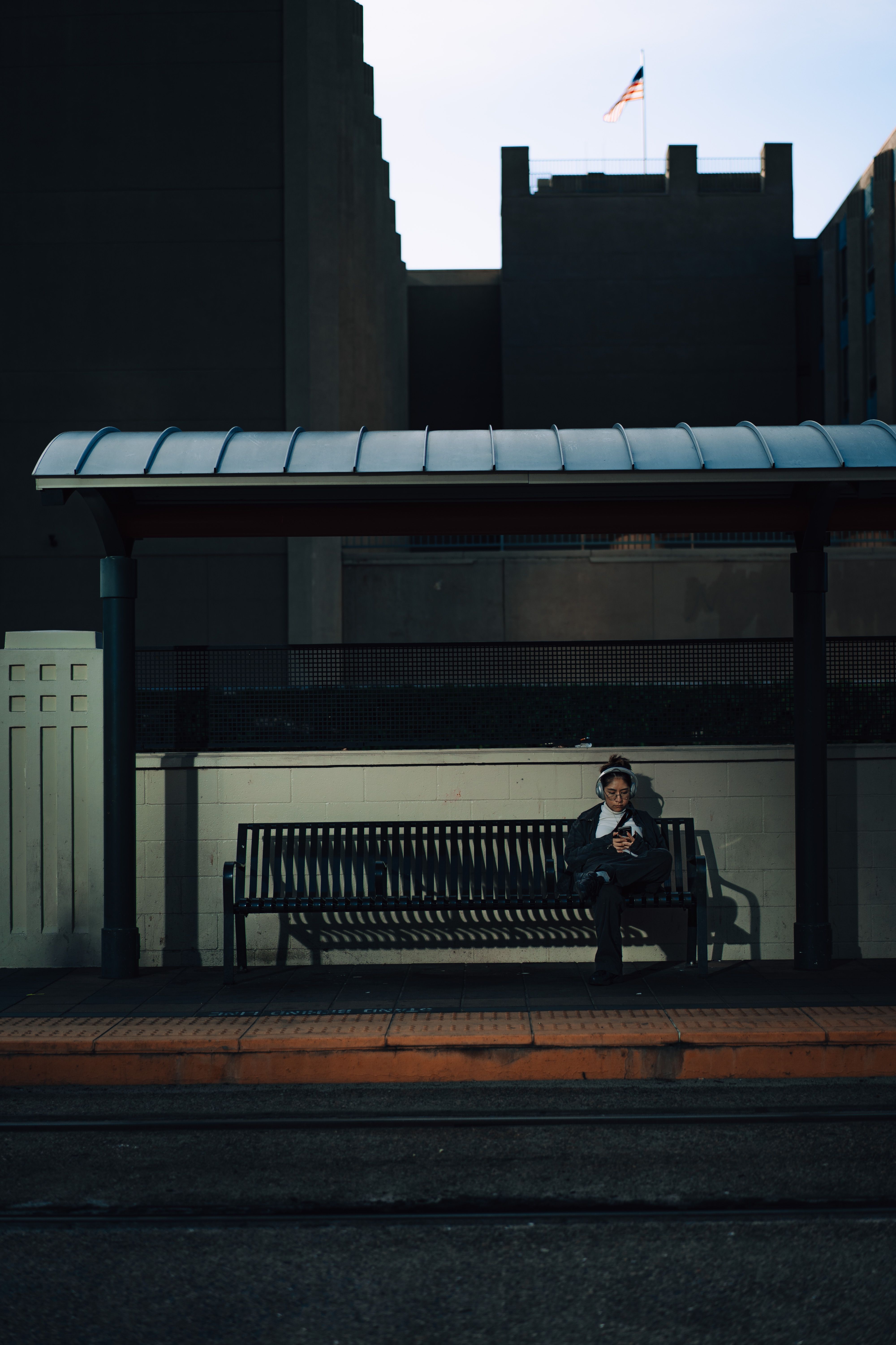 A solitary person sits at an urban tram stop bench during twilight, surrounded by buildings.