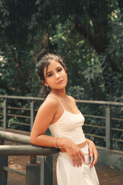 Free Stylish young woman in white outfit leaning on a handrail with greenery background. Stock Photo