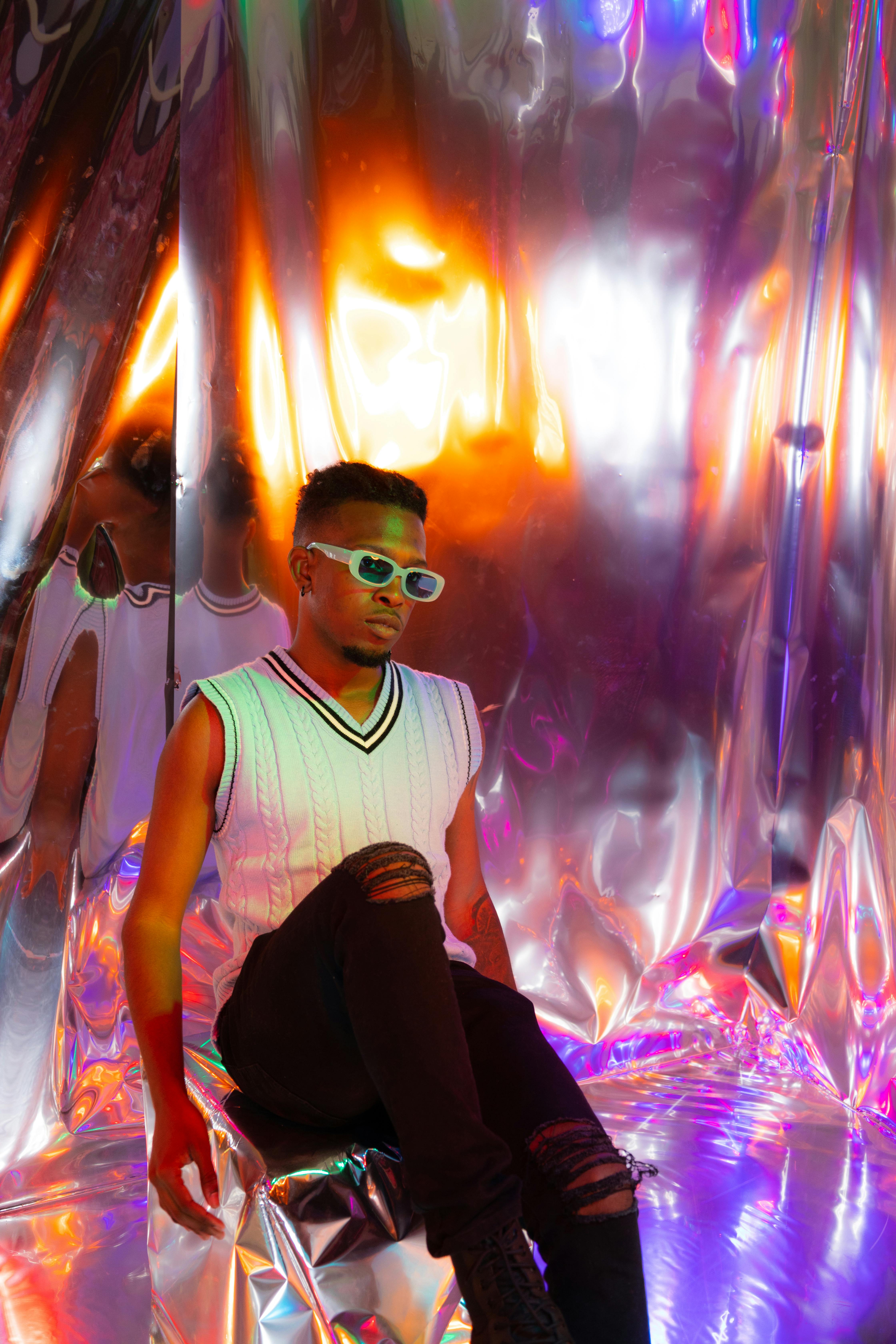 Fashionable man in sunglasses and sleeveless shirt in a reflective, colorful setting.