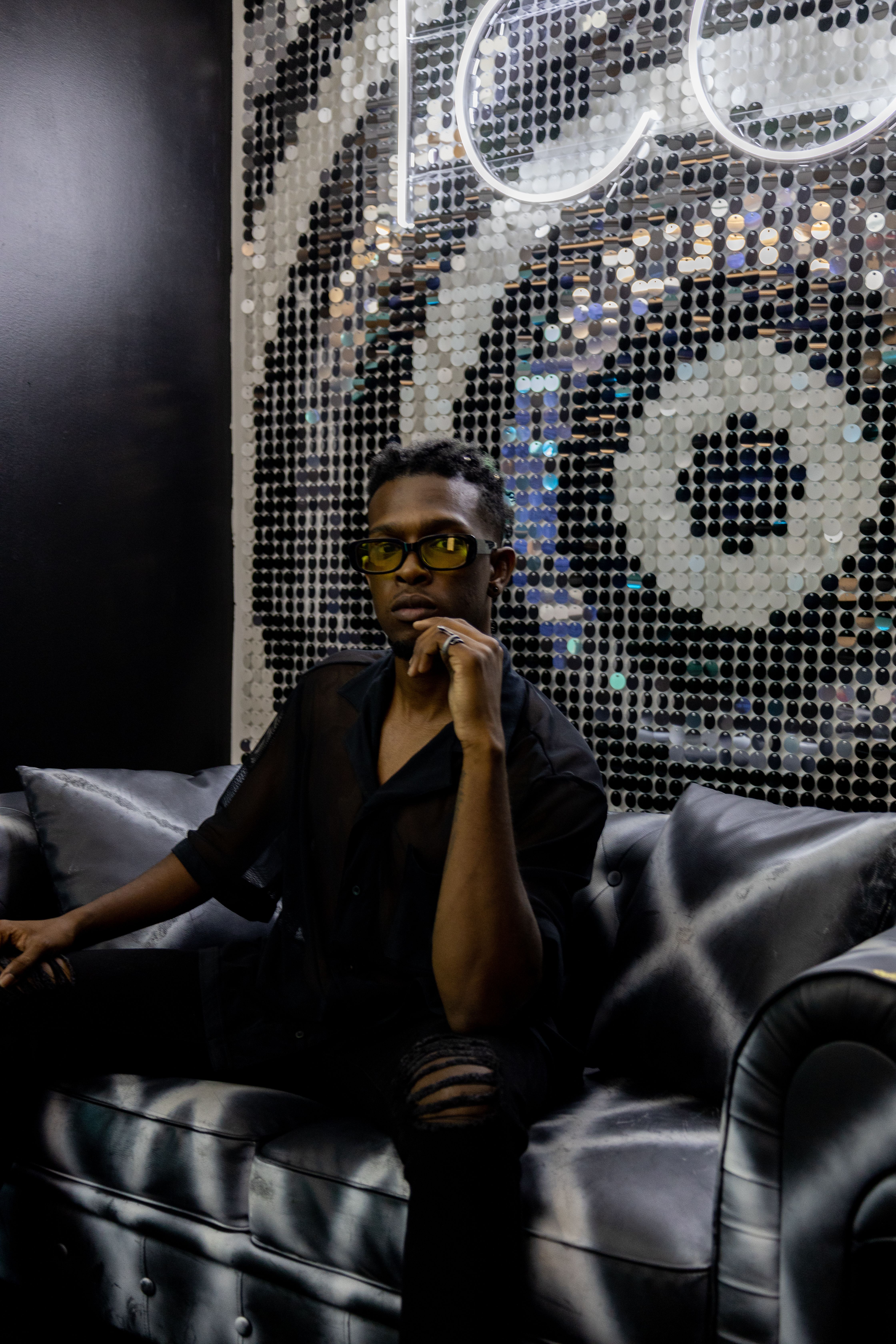 Sophisticated man wearing yellow sunglasses sitting on a leather couch against a shiny backdrop.