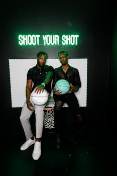 Two young men holding basketballs under neon sign 'Shoot Your Shot'.