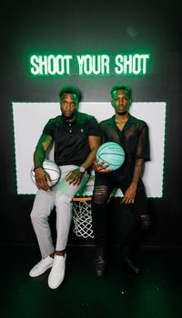 Two stylish men pose with basketballs under a neon sign, blending fashion and sports.