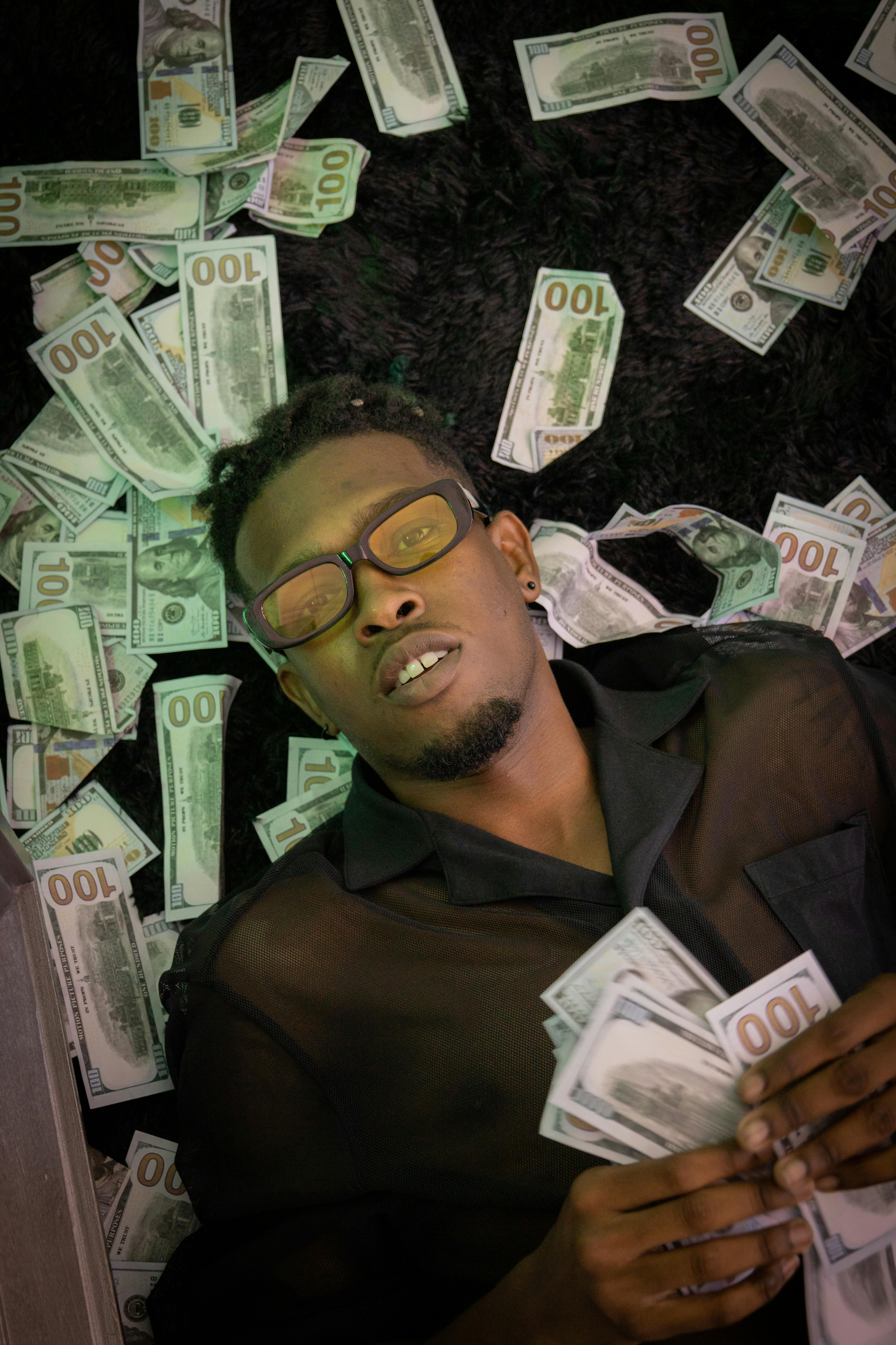 A young man with glasses lying on a pile of banknotes, embracing wealth and relaxation.