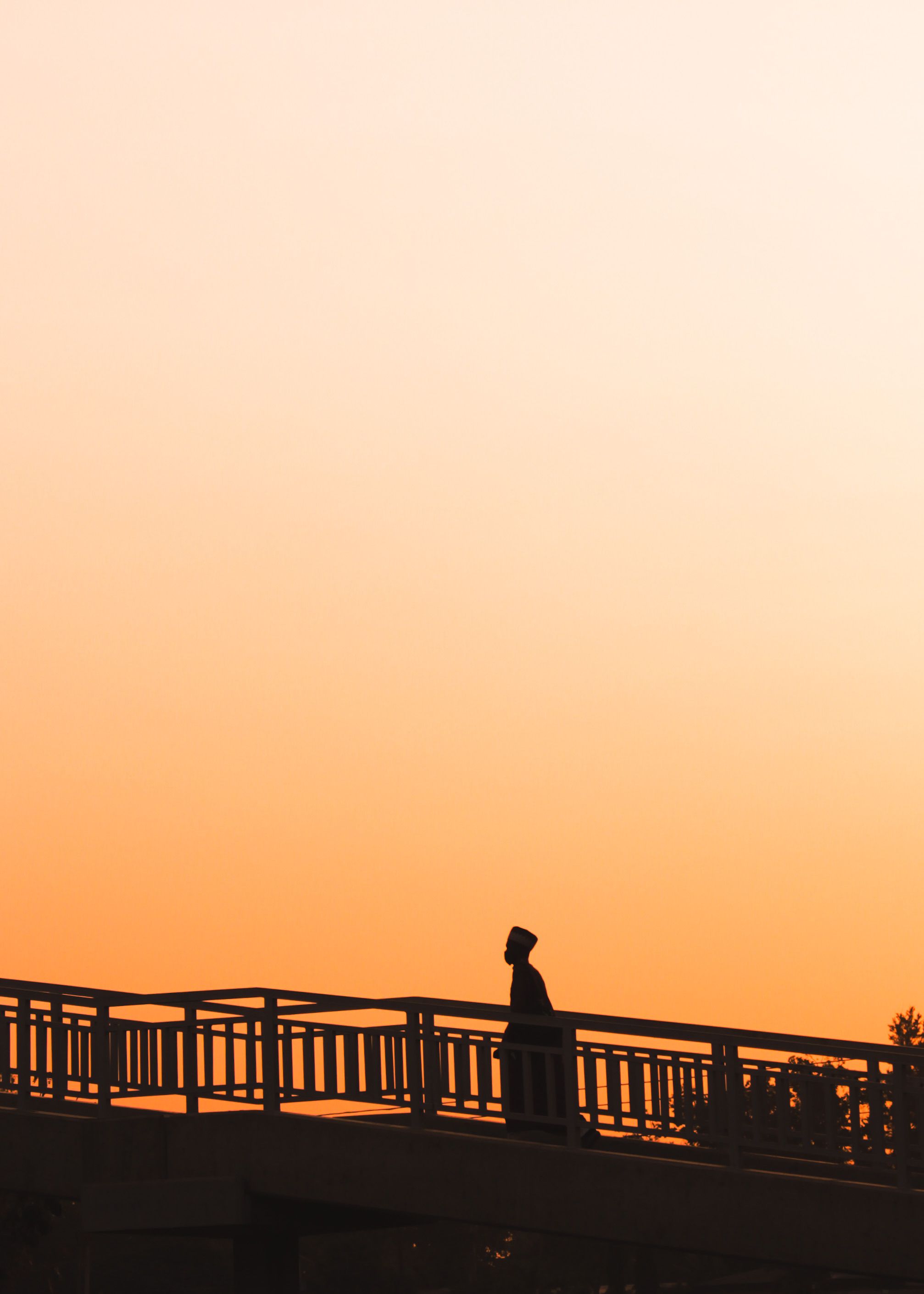A dramatic silhouette of a person walking on a bridge against an orange sunset sky in Kaduna, Nigeria.. pexels, dramatic, sil
